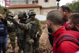 L'image montre une scène de tension avec des soldats en uniforme armés qui interagissent avec des civils. L'un des soldats, au premier plan, semble donner des instructions, tandis que d'autres soldats et des personnes en civil, certains portant des vêtements de protection, entourent la zone. L'environnement est urbain, avec des bâtiments en arrière-plan.