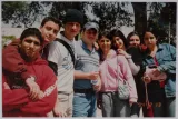 L'image montre un groupe de jeunes adultes posant ensemble en extérieur. Ils sourient et semblent être dans un endroit ensoleillé, entourés de quelques arbres. Les membres du groupe portent des vêtements décontractés, incluant des pulls et des t-shirts. Certains tiennent des bouteilles d'eau. L'ambiance semble joyeuse et amicale.