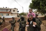 L'image montre un militaire portant une petite fille sur ses épaules, tandis que d'autres soldats armés avancent à proximité, sous un ciel nuageux et des bâtiments en arrière-plan.