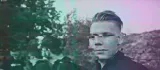 Un jeune homme au visage partiellement flou, entouré d'arbres et de personnes indistinctes.