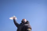 Un homme en manteau noir lève le bras vers le ciel, tenant une colombe blanche. Le fond est un ciel bleu clair.