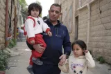 L'image montre un homme tenant un jeune enfant dans ses bras, tandis qu'il tient la main d'une petite fille à côté de lui. Ils se trouvent dans une ruelle étroite avec des bâtiments en pierre. L'homme porte une tenue de sport, et les enfants sont vêtus de vêtements colorés.