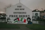 L'image montre un mur d'enceinte avec une inscription en anglais : "YOU ARE NOW ENTERING FREE DERRY". Le fond est blanc. Il y a également des graffitis artistiques représentant un enfant et un homme en uniforme avec un béret, portant un masque à gaz, ainsi qu'un drapeau. Les mots "RESISTANCE IS NOT TERRORISM" et "Saorise Don Phalastin" (qui signifie "Liberté pour la Palestine" en irlandais) sont également visibles. L'ensemble évoque un message politique fort lié à la lutte pour la liberté et la résistance.