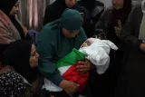 L'image montre une femme en deuil tenant un enfant enroulé dans un drapeau palestinien. Elle est entourée d'autres femmes, également en deuil, qui montrent une atmosphère de tristesse et de solidarité.