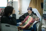 L'image montre un homme assis dans un fauteuil d'hôpital, portant un foulard coloré sur la tête. Son expression est sérieuse et il semble être dans un environnement médical, probablement en train de recevoir un traitement. Il est entouré d'autres personnes dans des sièges similaires. L'ambiance est calme, avec un éclairage doux provenant des fenêtres. L'homme est couvert d'une couverture avec un motif floral.