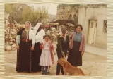 L'image montre un groupe de cinq femmes et une jeune fille, se tenant ensemble à l'extérieur. Elles sont entourées d'un paysage rural, avec des structures en arrière-plan, peut-être des maisons ou des granges. Les femmes portent des vêtements traditionnels, et la jeune fille est habillée d'une robe rose. Un chien se trouve également près d'elles. L'atmosphère de la photo semble conviviale et chaleureuse.