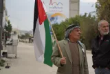 L'image montre deux hommes dans une rue. L'un d'eux, portant une tenue traditionnelle, tient un drapeau palestinien. L'autre homme, en veste noire, regarde en direction opposée. En arrière-plan, on aperçoit des bâtiments et de la végétation.