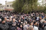 L'image montre une foule dense de personnes rassemblées dans une rue, avec des bâtiments en arrière-plan. Les gens semblent concentrés et attentifs, certains portant des vêtements sombres. Il y a des arbres et des feuillages, et l'atmosphère est celle d'un événement significatif.