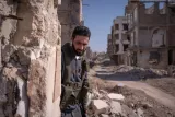 L'image montre un homme en tenue militaire, appuyé contre un bâtiment en ruines. Le paysage est dévasté, avec des structures détruites et des débris partout, sous un ciel dégagé. L'ambiance semble tendue et tragique.