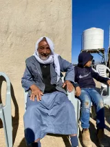 Sur cette image, on voit un homme âgé assis sur une chaise en plastique. Il porte un vêtement traditionnel et un foulard sur la tête. Son visage affiche un sourire chaleureux. À côté de lui, un jeune garçon portant un sweat à capuche et des jeans regarde vers le bas. En arrière-plan, on aperçoit un mur et un réservoir d'eau. Le ciel est dégagé, ce qui donne une atmosphère ensoleillée à la scène.