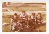 L'image montre un groupe de cinq jeunes hommes assis sur l'herbe. Ils portent tous des écharpes blanches et rouges enroulées autour de leur cou. L'ambiance semble décontractée et amicale. Au fond, on peut voir un paysage naturel avec un sol herbeux et des éléments architecturaux. Les expressions des jeunes sont souriantes, ce qui suggère qu'ils passent un bon moment ensemble.