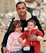 Un homme souriant tient un bébé, assis avec une petite fille dans un environnement en débris.