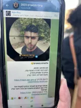 L'image montre un téléphone portable tenant une photo d'un jeune homme. L'écran affiche un message en hébreu avec des informations accompagnées d'une photo de la personne. Le tout semble contenir des détails sur un événement ou une publication, incluant un lien. En arrière-plan, on aperçoit quelques personnes floues, probablement dans un environnement extérieur.