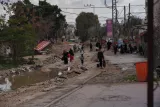 L'image montre une rue endommagée, avec des personnes marchant sur un sol irrégulier. Il y a de l'eau stagnante sur le côté, des bâtiments et des arbres en arrière-plan, ainsi que des panneaux et des fils électriques visibles. L'atmosphère semble sombre et désordonnée.