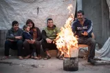 L'image montre un groupe de cinq personnes assises autour d'un feu. Ils semblent passer un moment convivial, avec des sourires et des expressions détendues. Le feu, qui brûle dans un bidon, crée une ambiance chaleureuse. En arrière-plan, il y a une tente ou une couverture qui sert de toile de fond. Les membres du groupe portent des vêtements décontractés. L'atmosphère semble informelle et accueillante.