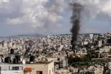 L'image montre une ville densément peuplée avec des bâtiments serrés. Dans le ciel, on voit de la fumée noire s'élever, indiquant une situation de conflit ou de détresse. Les nuages sont présents au-dessus, et le paysage urbain s'étend sur une colline.