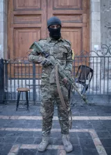 L'image montre un soldat portant un uniforme militaire camouflage, une cagoule et tenant un fusil. Il se tient debout devant un bâtiment avec une porte en bois. Un siège en bois est visible à l'arrière-plan.