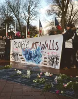 L'image montre une manifestation avec des personnes tenant une grande bannière sur laquelle est écrit "People Not Walls". Cette bannière est décorée de dessins, notamment une illustration symbolique. Au sol, on aperçoit des fleurs, probablement en hommage aux victimes. En arrière-plan, des drapeaux français sont visibles, et l'atmosphère semble réfléchir un message de solidarité et d'humanisme. Le rassemblement se déroule dans un espace public, suggérant un engagement pour une cause sociale.