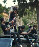 L'image montre un groupe de quatre personnes en pleine performance artistique sur le toit d'une voiture. Ils portent tous des vêtements noirs. Deux d'entre eux semblent jouer des instruments, tandis que les autres se déplacent de manière dynamique. L'environnement est urbain, avec des arbres et des bâtiments en arrière-plan, ce qui ajoute une ambiance vivante à la scène. L'énergie de la performance est palpable, suggérant une interaction ludique entre les participants.