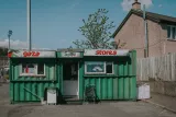 L'image montre un petit magasin qui semble être un commerce de proximité. Le bâtiment est en conteneur maritime peint en vert, avec des inscriptions "gaza" et "stores" sur le dessus. Il y a des fenêtres sur le devant avec des affichages visibles à l'intérieur. À côté du magasin, il y a un petit fauteuil, et l'arrière-plan montre des maisons et un ciel partiellement nuageux. L'ensemble de la scène donne une impression de quartier calme et modeste.