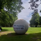 L'image montre une grande sphère blanche située dans un espace vert, probablement un parc. Sur la sphère, il y a une banderole avec le message "FREE PALESTINE", ainsi qu'une mention de "STUDENTS FOR PALESTINE." L'environnement est paisible, avec des arbres et un ciel nuageux en arrière-plan.