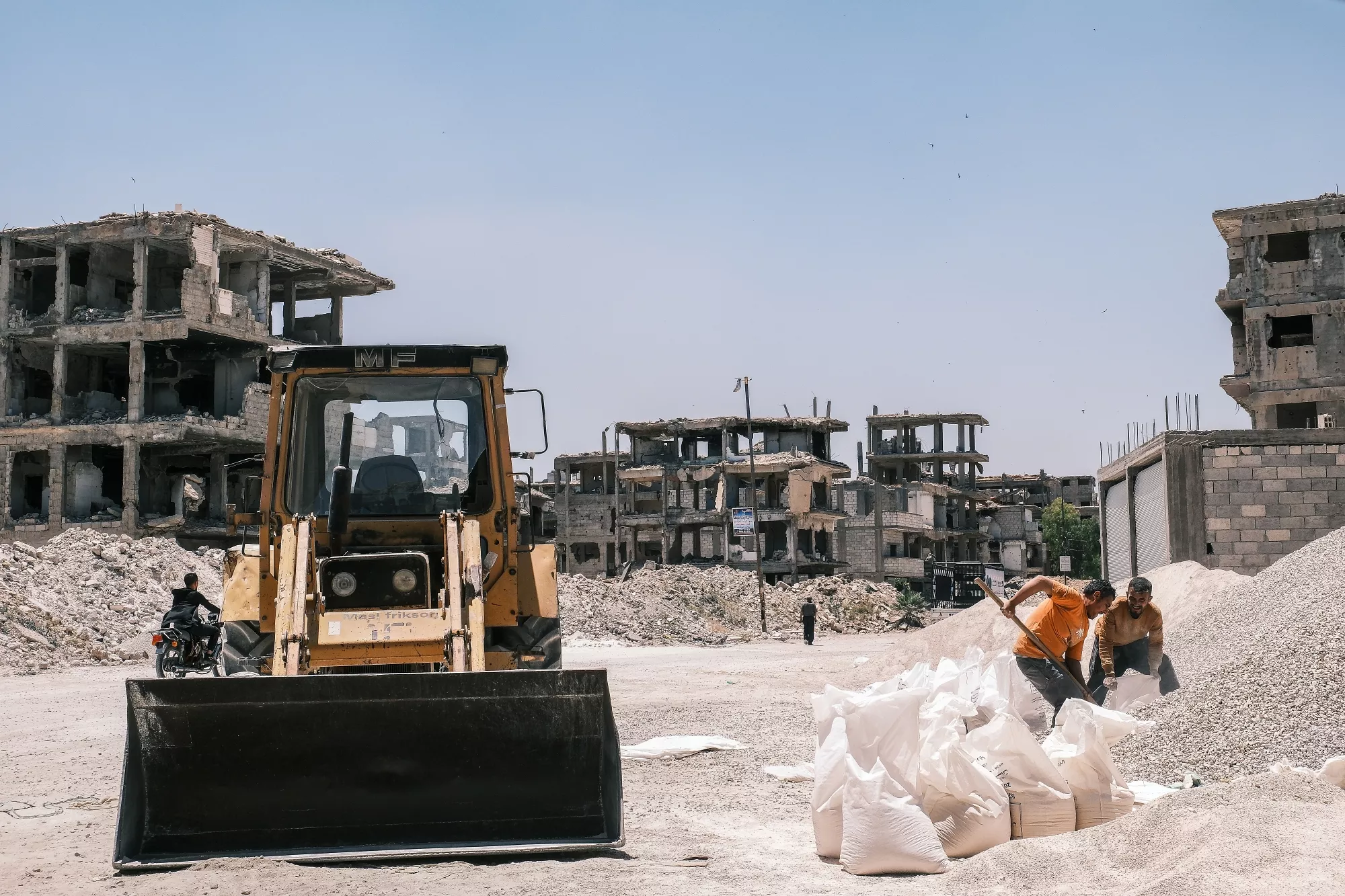 Terreno devastato con rovine e operai al lavoro in un cantiere di ricostruzione.