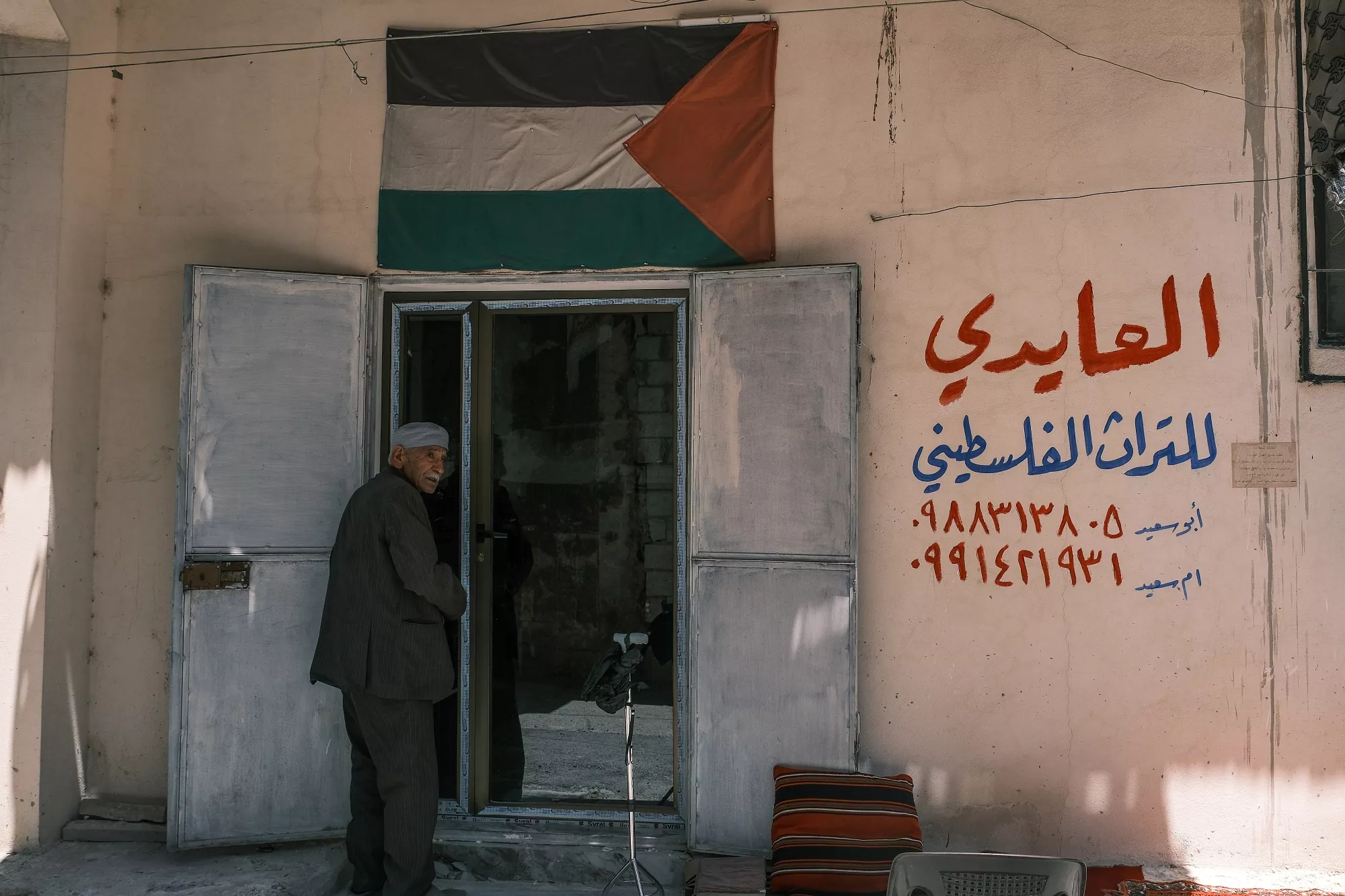 Un uomo anziano è in piedi davanti a una porta, con in mano una bandiera palestinese e una scritta in arabo.