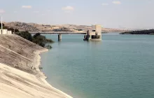 تظهر الصورة منظرًا هادئًا لبحيرة محاطة بتضاريس جبلية. يتوسط المشهد بناء هندسي يُحتمل أن يكون محطة لتحكم في المياه أو وسيلة لتخزينها. المياه باللون الأزرق الفاتح تعكس السماء، بينما تمتد ضفاف البحيرة بلطف، مع وجود تدرجات من اللون الرمادي الفاتح في الرمال. الأفق يكتمل بكتل من التلال الخضراء البعيدة، مما يضفي على الصورة جمالًا طبيعيًا وهدوءً.
