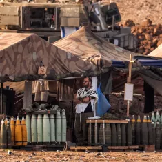 The image depicts a military encampment with tents set up in a rugged landscape. In the foreground, a soldier wearing a tallit (a traditional Jewish prayer shawl) stands near stacks of artillery shells arranged on a wooden platform. The tents are camouflaged, blending into the environment, and various military equipment is visible in the background. The setting conveys a sense of military readiness and activity.