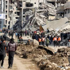 The image depicts a scene in Gaza featuring a street filled with people walking. In the background, there are buildings that appear to be damaged or destroyed. A large banner stretches across the street, welcoming visitors to Gaza, and some individuals wearing orange vests are visible, likely indicating they are part of a relief or aid organization. The area is marked by piles of debris and dirt, suggesting recent conflict or disaster. Overall, the atmosphere conveys a mix of resilience amid the challenges faced by the community.