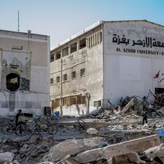 The image depicts a scene of devastation, likely in a war-torn area. In the foreground, there is rubble and debris from damaged buildings, suggesting destruction from conflict. A partially destroyed building is visible, with the name "Al Azhar University - Gaza" written on it in Arabic, indicating it is an educational institution. In the foreground, two individuals on bicycles navigate through the debris, highlighting the struggle of daily life in such an environment. The overall atmosphere conveys a sense of loss and destruction, with remnants of architecture marked by chaos.