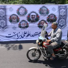 تصویر مردی سوار بر موتورسیکلت که در پیش‌زمینه، بنر بزرگ و مردم نشسته در پشت آن دیده می‌شوند.