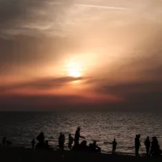 The image depicts a beach scene during sunset. In the foreground, there are silhouettes of people walking along the shore, some sitting or standing by the water. The sky is filled with soft hues of orange and pink, blending into darker clouds as the sun sets on the horizon. The water reflects the colors of the sky, creating a tranquil and serene atmosphere. Overall, the scene conveys a sense of calm and natural beauty as day transitions into night.