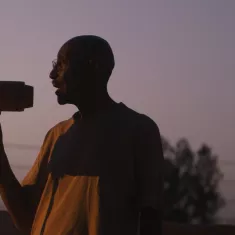 رجل يحمل مكبر صوت، silhouetted أمام غروب الشمس، يبدو وكأنه ينادي بشيء مهم.