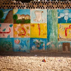 The image shows a wall decorated with colorful murals or paintings. The artwork includes various designs such as flowers, shapes, and possibly symbols or letters that are arranged in a grid pattern. The colors are vibrant, featuring shades of blue, green, yellow, and pink, giving the wall a lively and artistic feel. The surrounding area appears dusty or sandy, suggesting an outdoor environment. Overall, the scene conveys a sense of creativity and community.