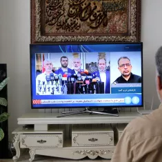 A person is sitting on a couch, facing a television that is displaying a news broadcast featuring several speakers at a press conference. In the background, there is a decorative wall hanging and a plant to the side.