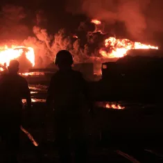The image depicts two silhouetted figures walking towards a large fire at night, with bright flames and thick smoke rising in the background. The scene conveys a sense of danger and urgency.