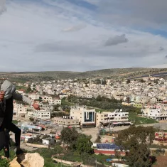 Nell'immagine si vede un giovane che si sta arrampicando su una roccia, con una città sullo sfondo. La città è composta da edifici di diversi piani, molti dei quali di colore chiaro. L'area è circondata da colline verdi e il cielo è parzialmente nuvoloso, suggerendo un clima variabile. Si possono notare anche alcune strutture agricole e aree di terreno aperto in primo piano.