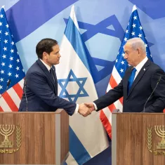 The image shows two men shaking hands at a press conference. They are standing at podiums, with Israeli and American flags in the background.