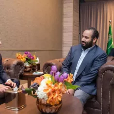 دو مرد در یک اتاق مرتب نشسته‌اند. یکی با کتی تیره و دیگری با پیراهن روشن. گل‌های رنگارنگ روی میز است.