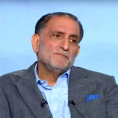 Hombre de mediana edad con barba, vestido formalmente, está pensando y mirando hacia un lado. Fondo desenfocado.