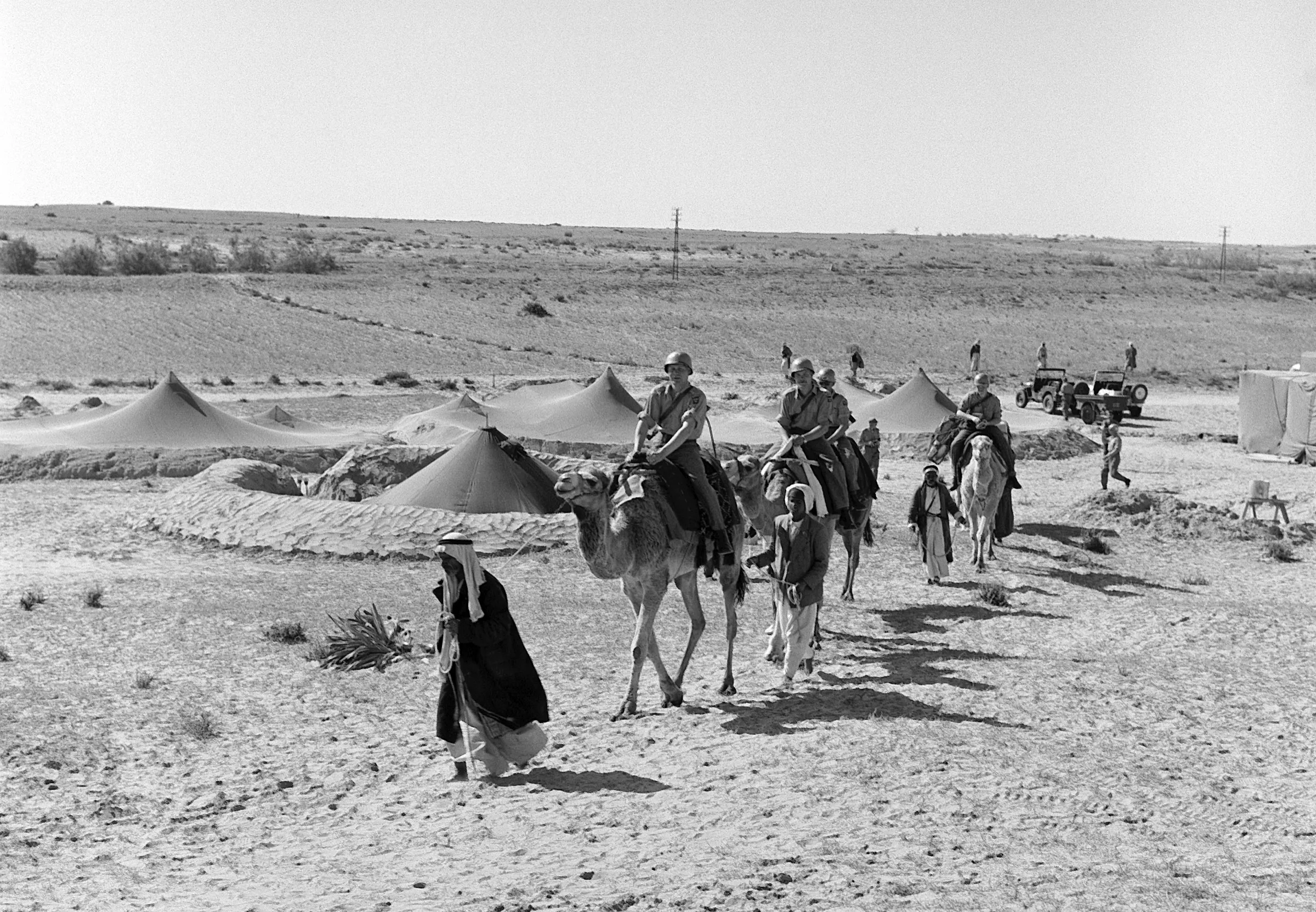 Un groupe de personnes traverse le désert à dos de chameau, près de tentes.