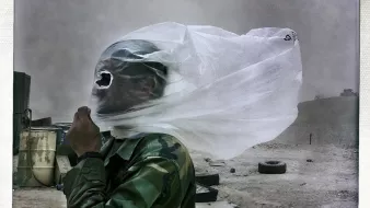 The image depicts a person in military attire, seemingly in a harsh, dusty environment. Their face is partially obscured by a white plastic sheet or bag that is being blown by the wind, creating a dramatic effect. The surroundings appear barren, with hints of debris and tires in the background, suggesting a desolate or rugged terrain. The overall atmosphere conveys a sense of struggle against the elements.