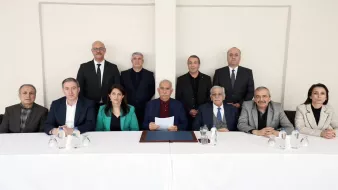 L'image montre un groupe de douze personnes assises derrière une table blanche. Il y a des hommes et des femmes, tous vêtus de costumes et de tenues professionnelles. L'ambiance semble formelle, et une personne au centre tient un document.