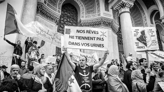 La imagen muestra una protesta o manifestación donde un grupo de personas sostiene pancartas y carteles. En el centro, un hombre sostiene un gran cartel con el mensaje "NOS RÊVES NE TIENNENT PAS DANS VOS URNES", que se traduce como "Nuestros sueños no caben en sus urnas". La escena está llena de gente, algunos con banderas y otros con rostros determinados, todos expresando descontento o reivindicaciones. El ambiente es de energía colectiva y compromiso social, y la imagen es en blanco y negro, lo que le da un tono más dramático y solemne.