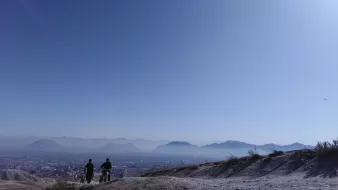 The image depicts a serene landscape with two individuals walking along a trail on a hillside. They are silhouetted against a vast, clear blue sky, which suggests a sunny day. In the background, there are distant mountains and a sprawling cityscape, partially visible below. The scene captures a sense of tranquility and openness, highlighting the natural beauty of the setting.