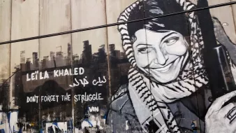 La imagen muestra un mural pintado en una pared, con un retrato en blanco y negro de una mujer que sonríe, vistiendo un keffiyeh. Junto a su imagen, hay texto en inglés que dice "DON'T FORGET THE STRUGGLE" y en árabe el nombre "ليلى خالد" (Leila Khaled). Este mural parece rendir homenaje a la figura de Leila Khaled, conocida por su activismo político y su papel en la lucha palestina. El fondo de la pared tiene grafitis y otros símbolos, lo que añade contexto urbano y de resistencia.