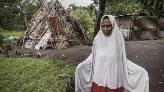 Sur cette image, on voit une femme debout au premier plan, portant un vêtement traditionnel avec un voile qui couvre sa tête. Son expression semble sérieuse ou réfléchie. À l'arrière-plan, on aperçoit une structure habitable, partiellement en bois et en matériaux naturels, qui semble être endommagée. La scène est entourée de verdure, avec des arbres et de l'herbe. L'ambiance générale évoque un contexte rural ou tribal, suggérant une connexion forte avec la nature et un mode de vie simple.