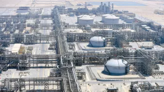 The image depicts a large industrial facility, likely an oil refinery or petrochemical plant. It features numerous large storage tanks, interconnected pipelines, and extensive infrastructure. The site is expansive, suggesting significant industrial activity, and it appears to be located in a relatively arid region, as indicated by the surrounding landscape. There are various structures for processing and storage, emphasizing the complexity and scale of the operation.