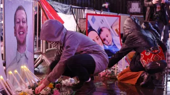 L'image montre des personnes en train de rendre hommage devant des portraits posés au sol. On peut voir des bougies allumées et des fleurs disposées autour des photos. Certaines personnes s'agenouillent ou se penchent pour arranger les fleurs. L'environnement semble humide, peut-être en raison de la pluie, et les participants portent des vêtements adaptés aux intempéries, comme des imperméables et des écharpes. L'atmosphère semble solennelle et empreinte de respect.